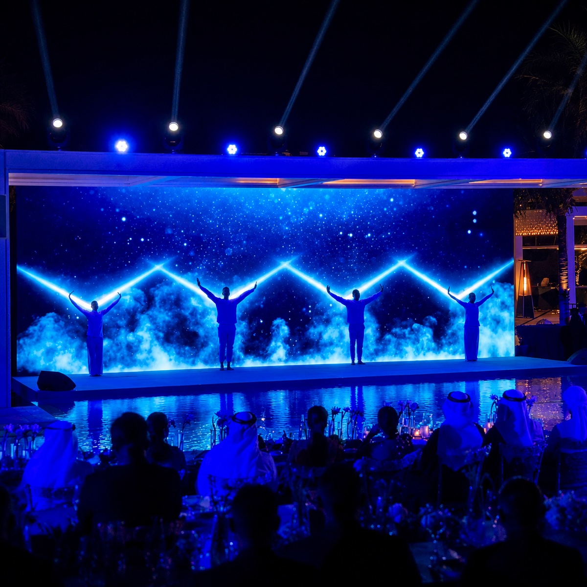 Four performers standing with arms raised on a poolside stage in front of a deep blue screen, guests seated at tables in front.