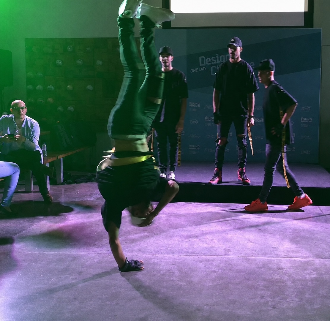 A dancer holding a one-handed handstand under green and magenta light, with other performers standing on a low stage behind.