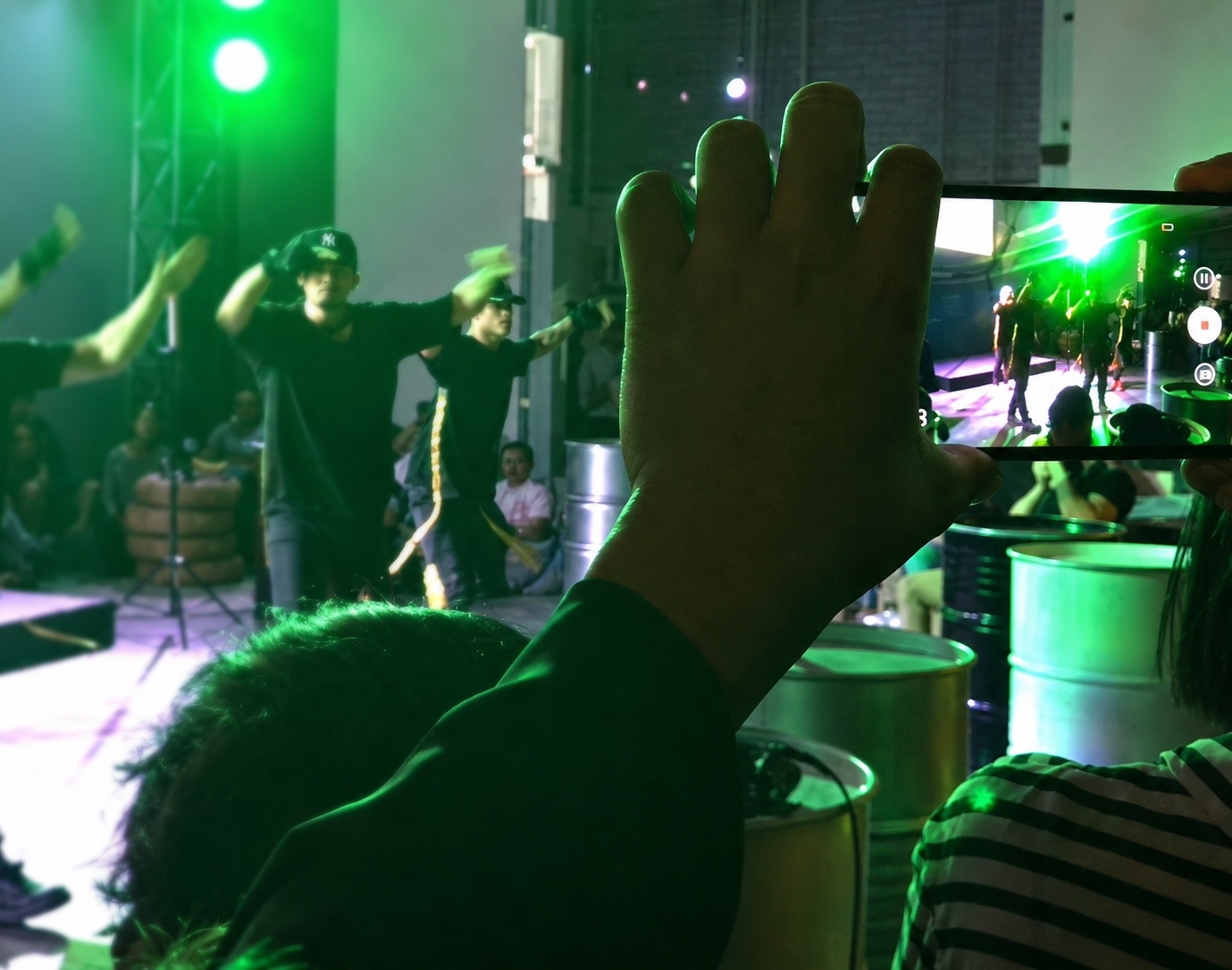 View from within the audience: a raised hand and a phone filming dancers performing under green light.