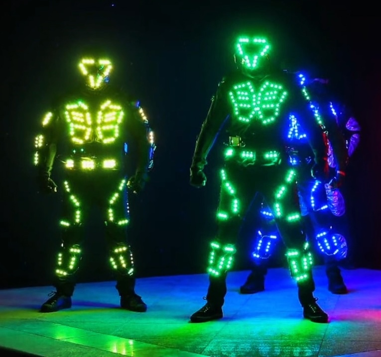 Two performers in LED costumes lit green and blue, standing on a small stage in a darkened room.