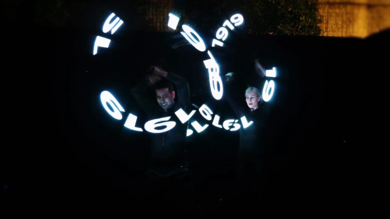 Long-exposure light trails as performers spin illuminated props, tracing the numbers 100 and 1919 in the air.