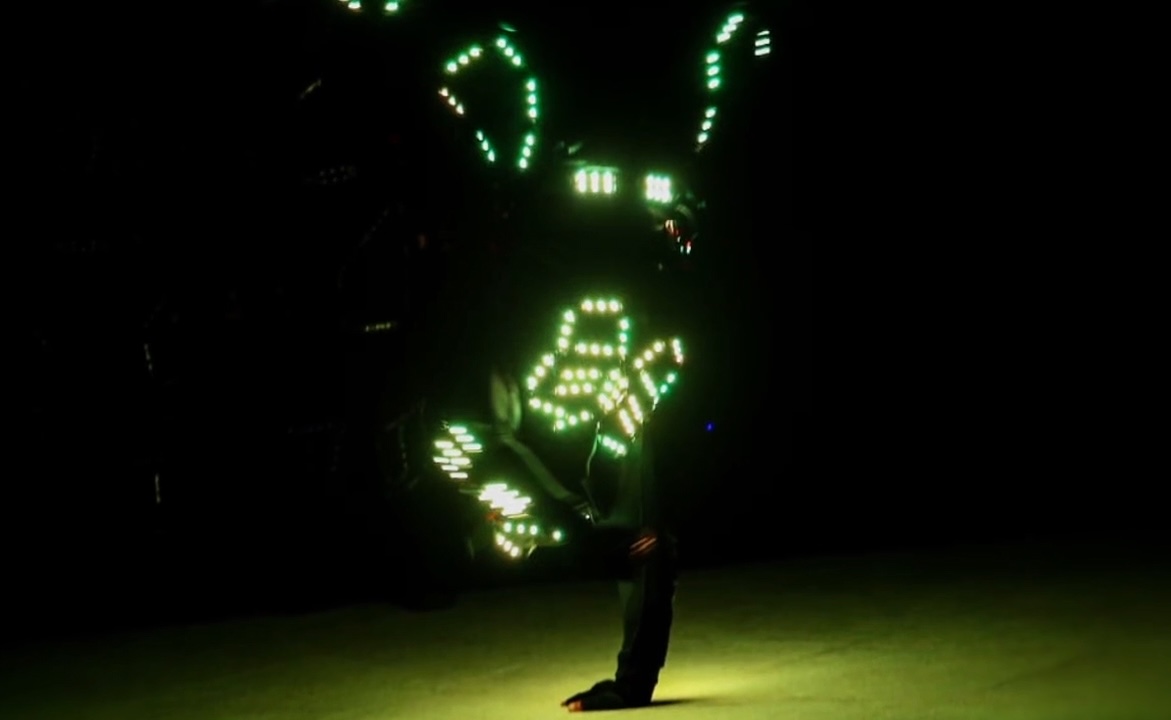 A single performer mid-move in near darkness, the green pixels of an LED costume tracing the body.