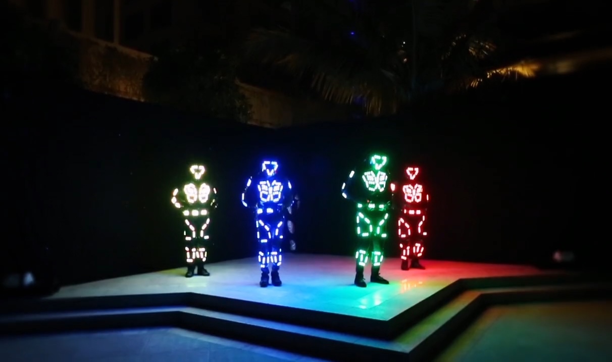 Four performers in LED costumes lit white, blue, green and red, standing on a poolside stage at night.
