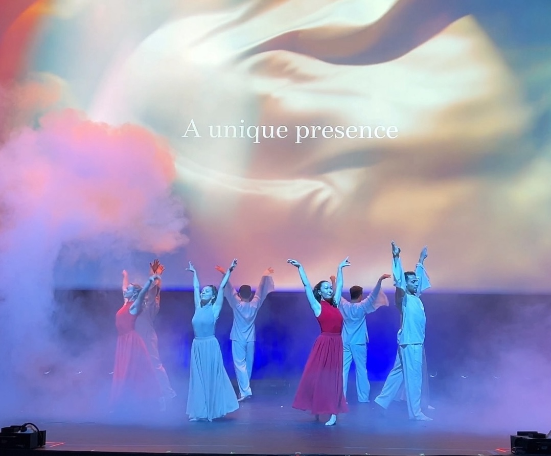 Dancers in pale flowing costume reaching upward through haze, in front of a screen reading “A unique presence”.