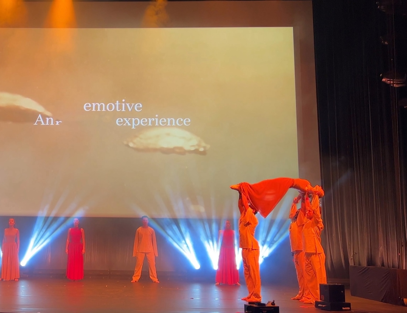 Performers in orange and red spread across a smoke-lit stage, two of them lifting a length of orange cloth overhead.
