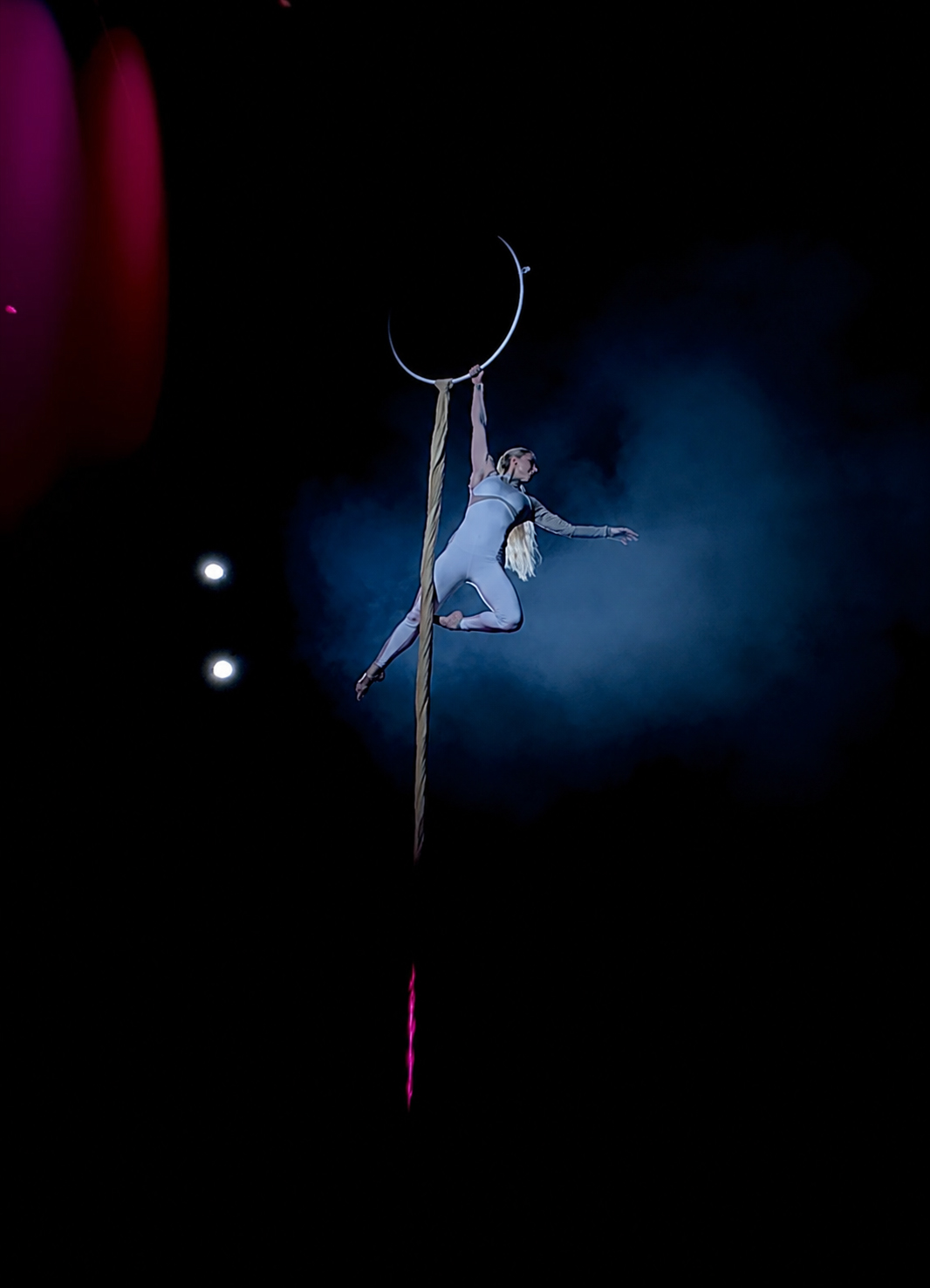 An aerial performer suspended on a vertical rope high above a darkened stage.