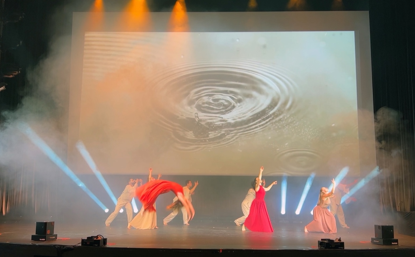 Dancers in red and cream mid-movement below a wide screen showing ripples spreading across water.