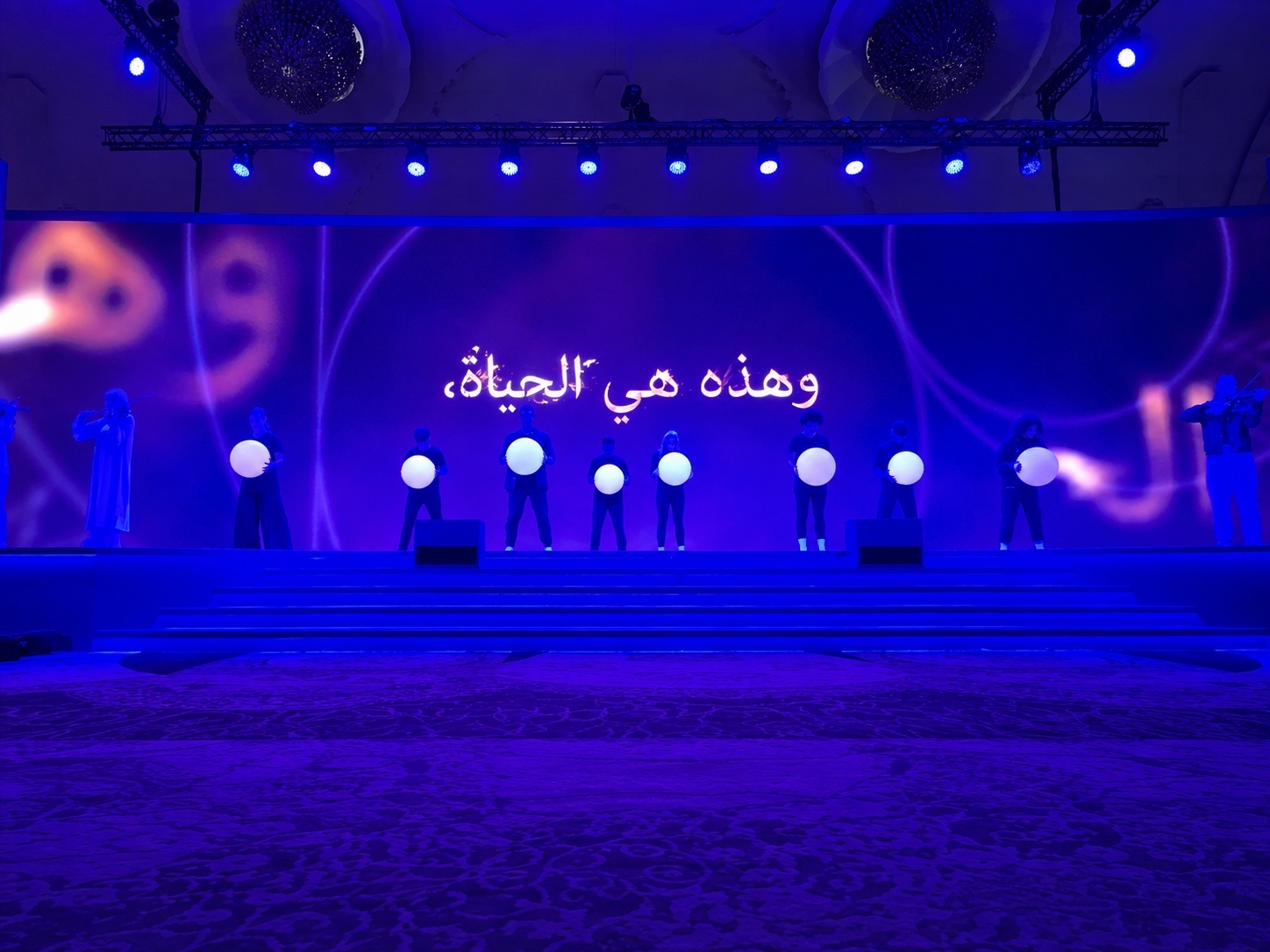 Performers holding illuminated white spheres across a blue-lit stage, Arabic lettering on the screen behind.