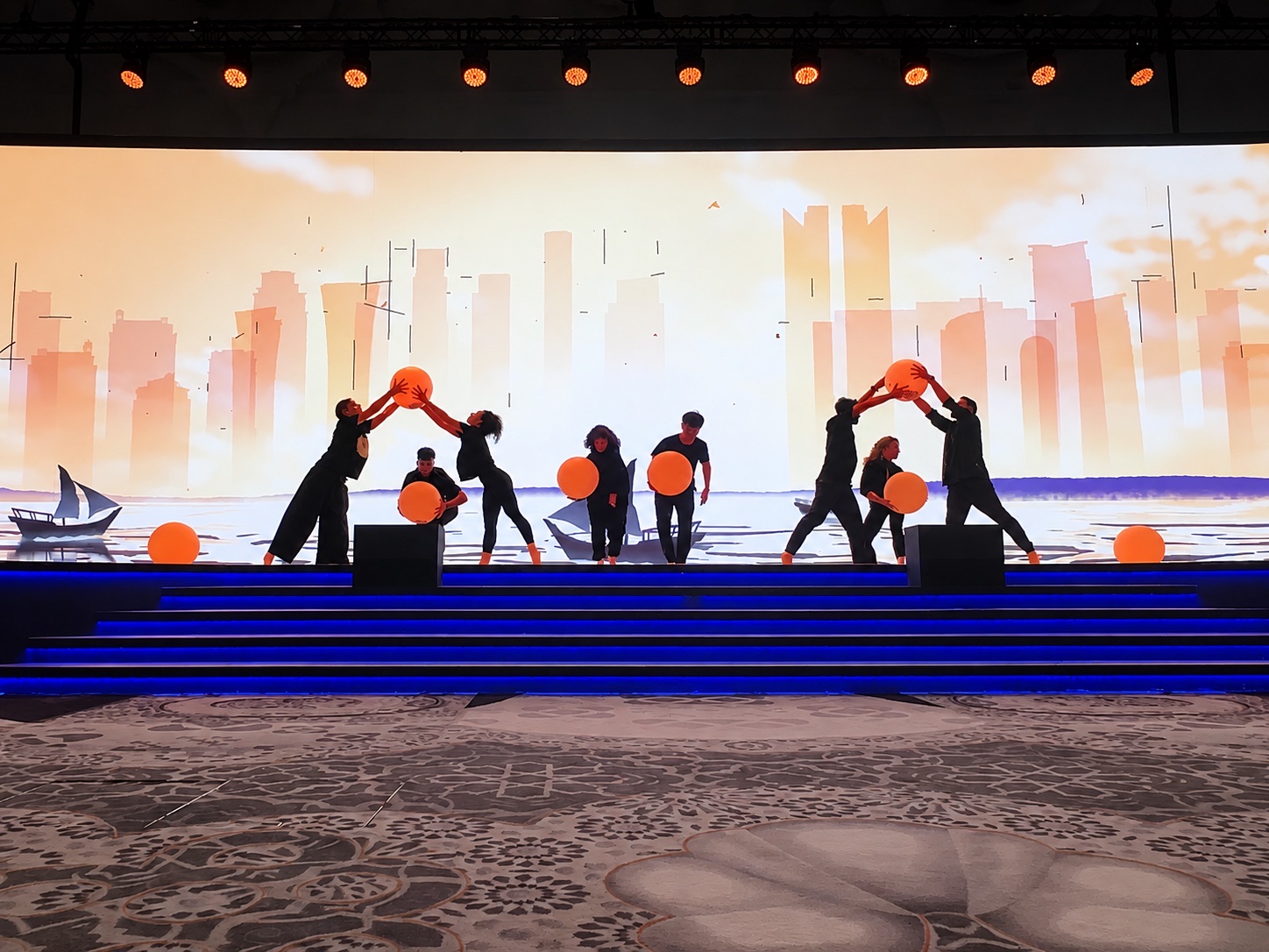 Performers silhouetted as they move large orange spheres in front of an illustrated city skyline.