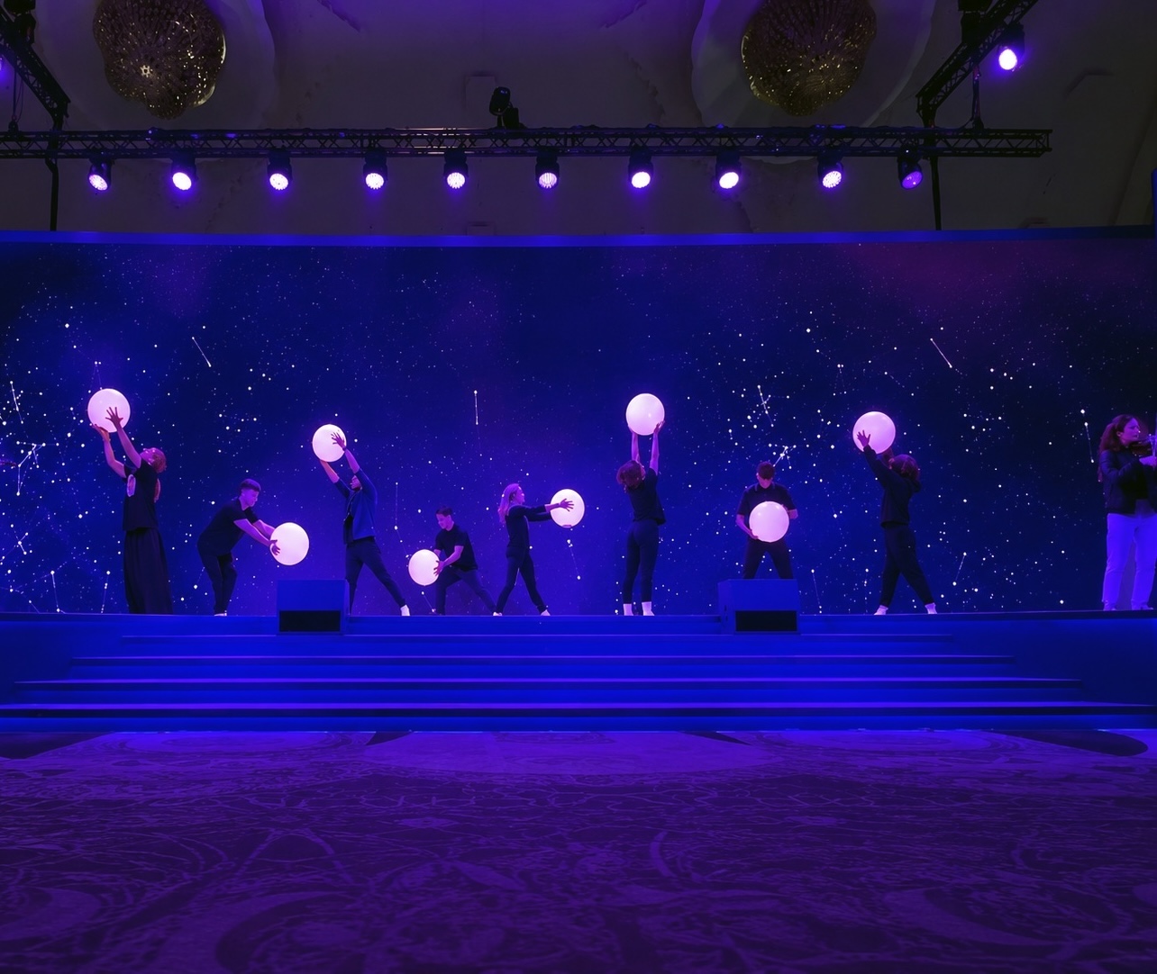 Performers spread across a stage holding glowing spheres against a deep blue starfield backdrop.