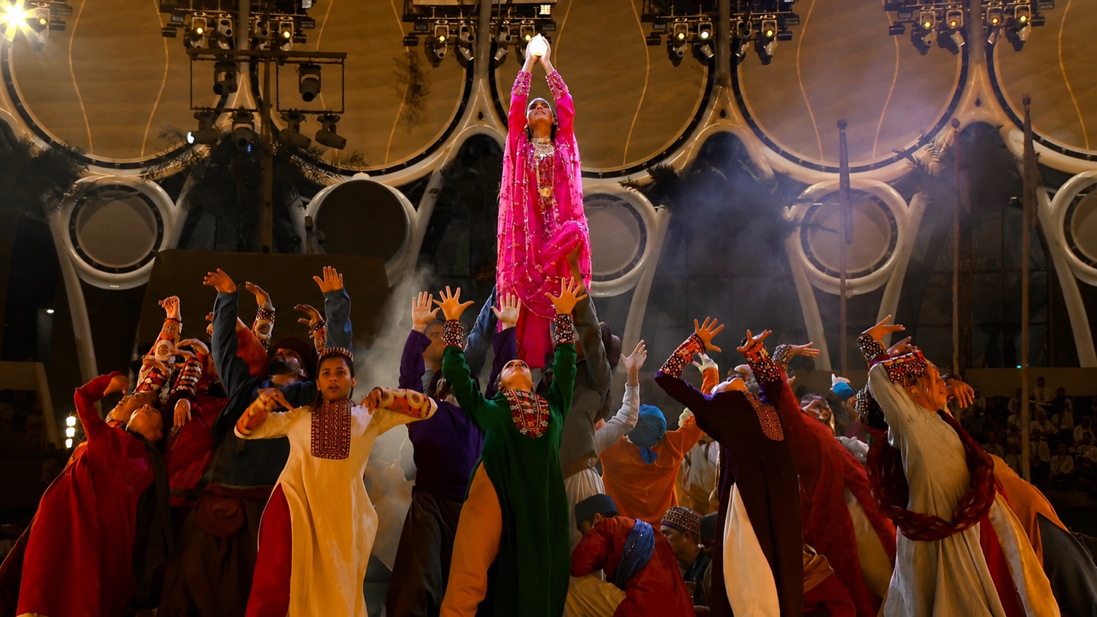 A performer in a pink robe raised above a crowd of costumed performers reaching upward beneath a domed roof.