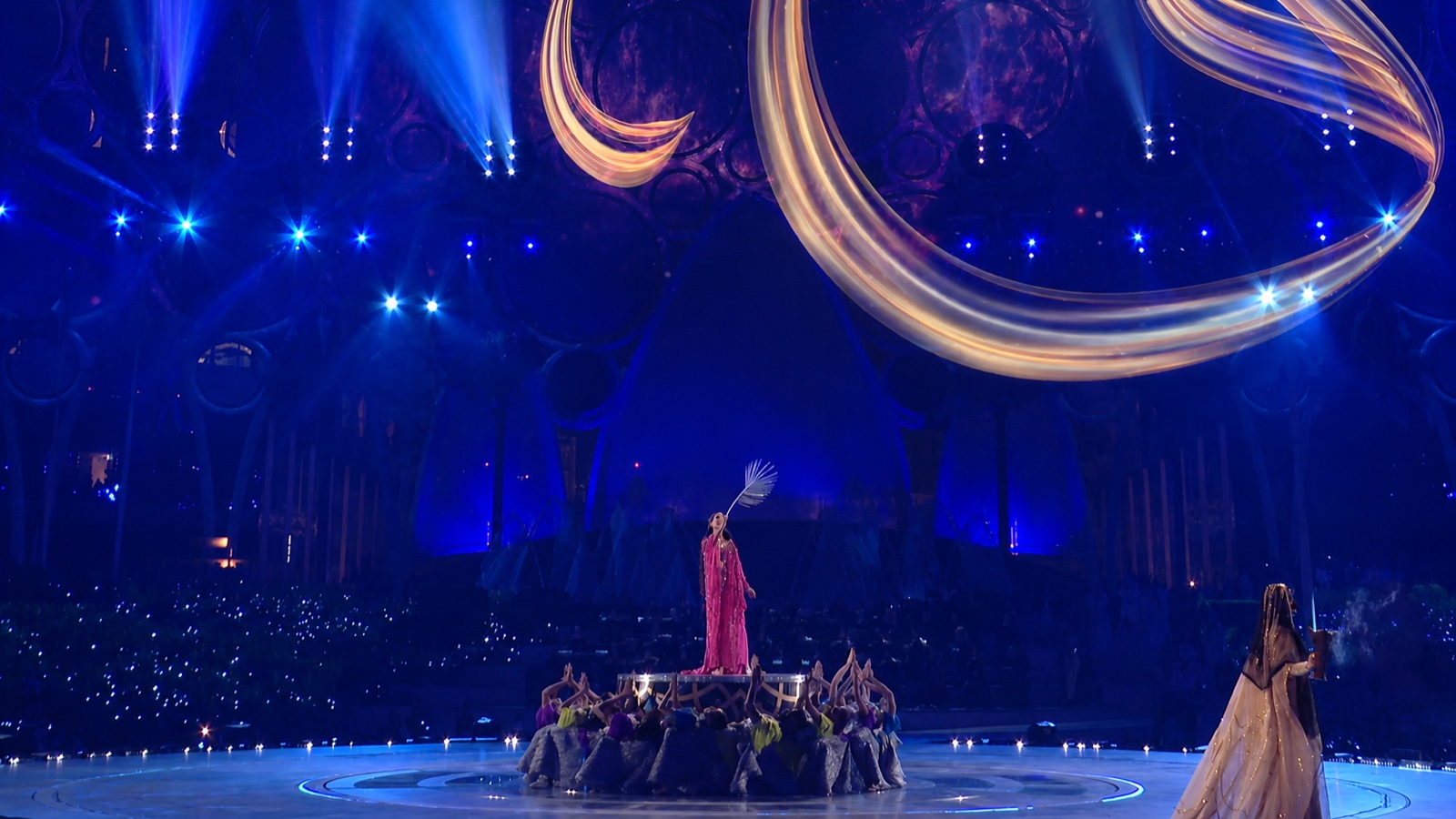 Wide view of the domed venue: a soloist on a raised platform as golden ribbons of projection sweep across the dome.
