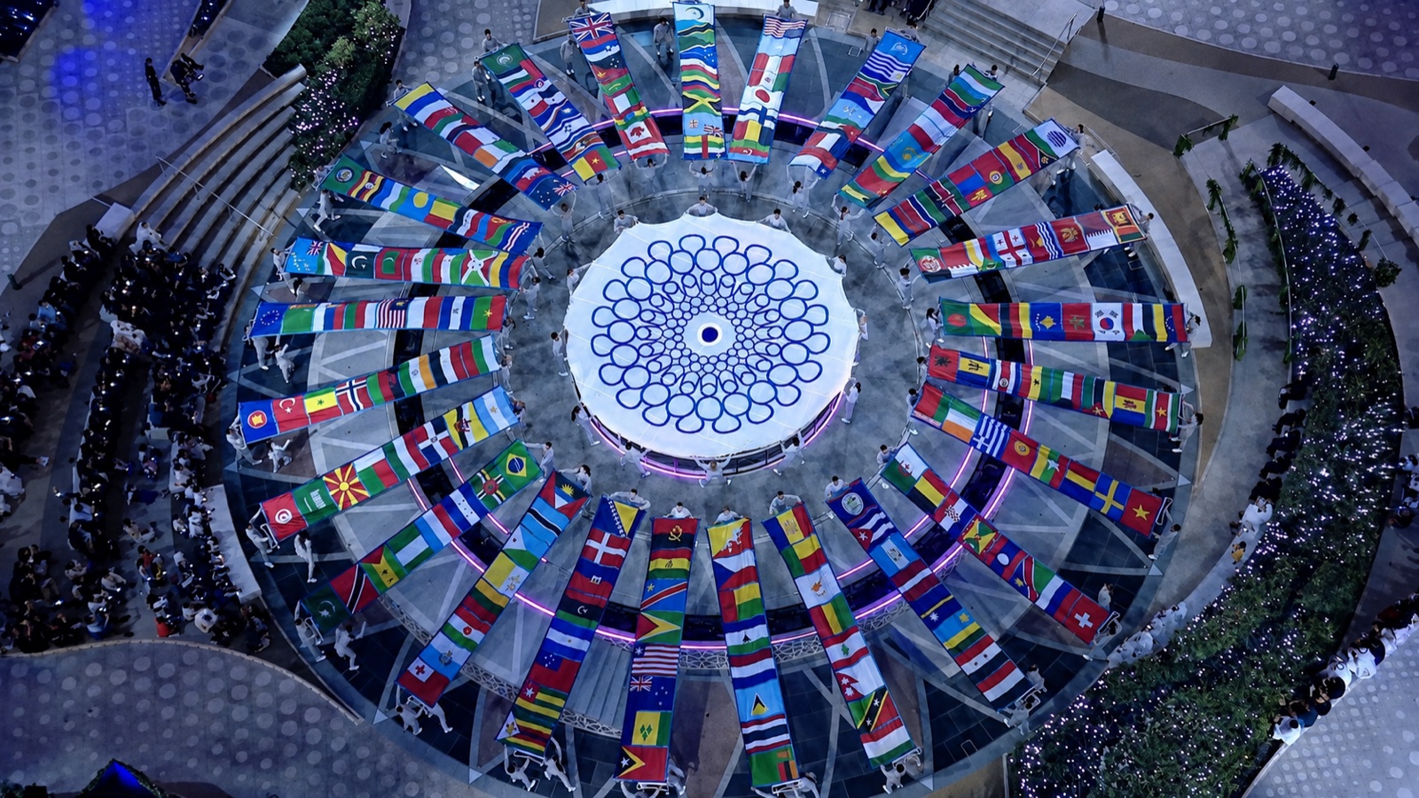 Aerial view of the circular plaza ringed by national flags, with the audience seated around it.