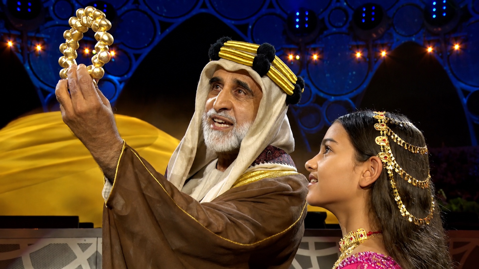 A performer in a white robe and headdress holds up a gold ornament beside a performer in embroidered dress.