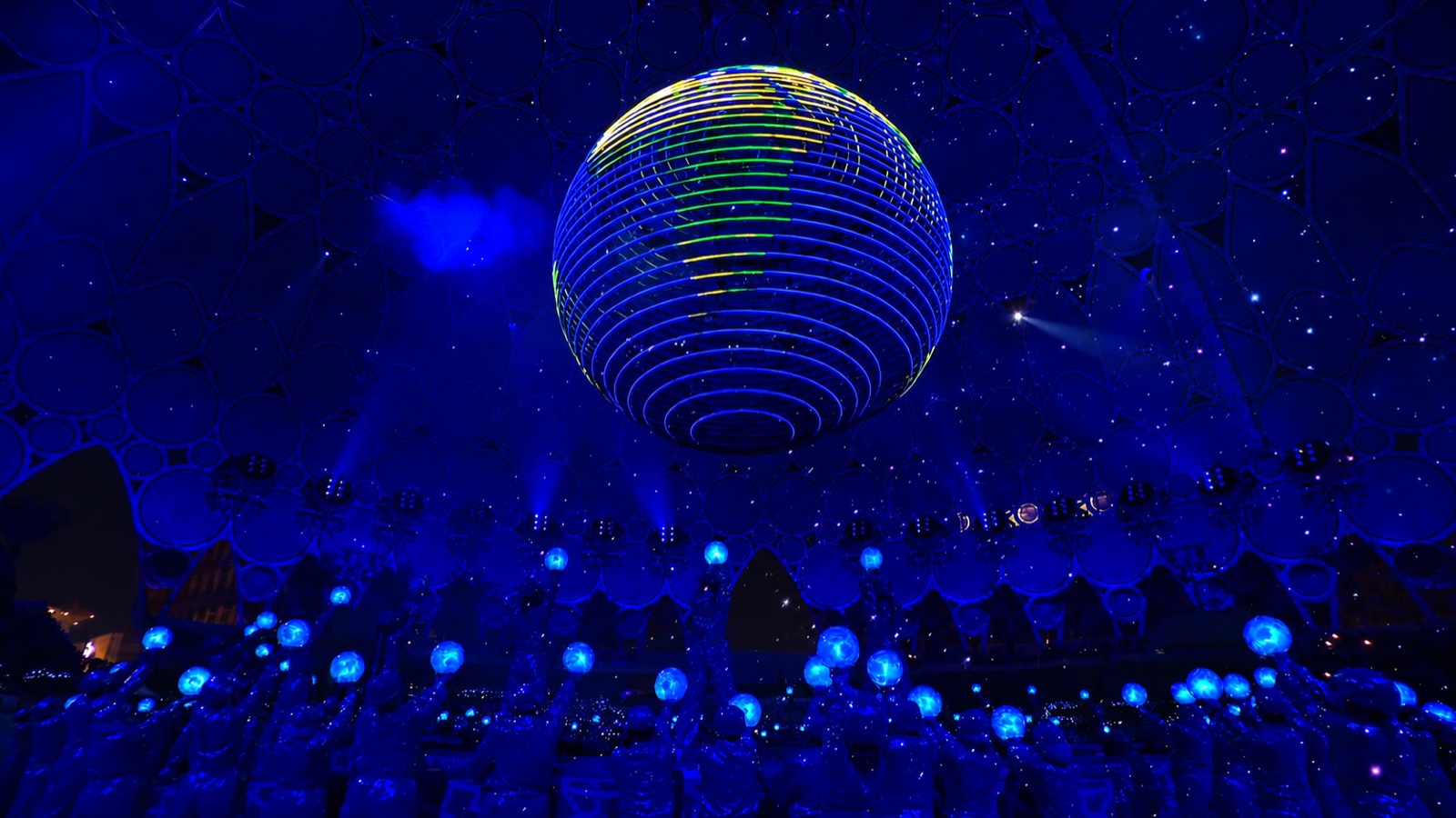 A large illuminated globe suspended above an audience holding glowing spheres, the room lit deep blue.