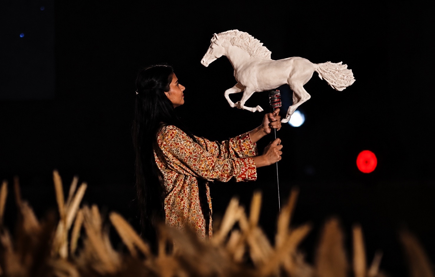 A performer holding a white horse puppet above a field of wheat-coloured props.