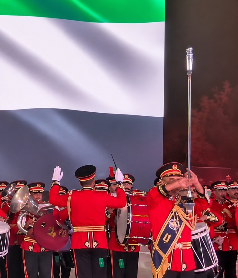 A marching band in red ceremonial uniform playing in front of a large projected UAE flag.