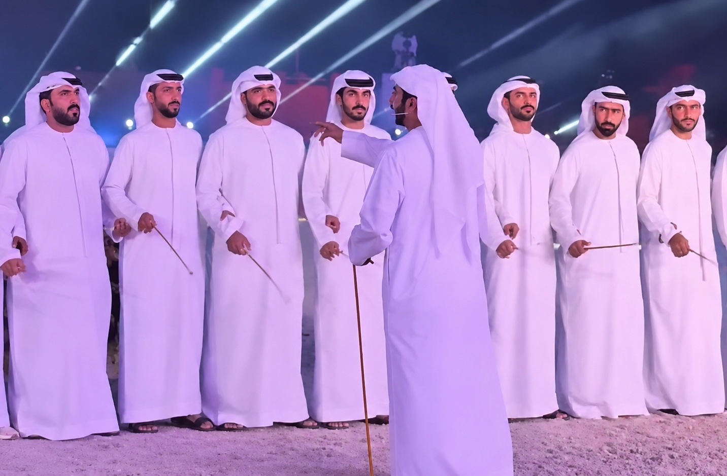 A line of performers in white robes holding thin canes in a traditional group dance.