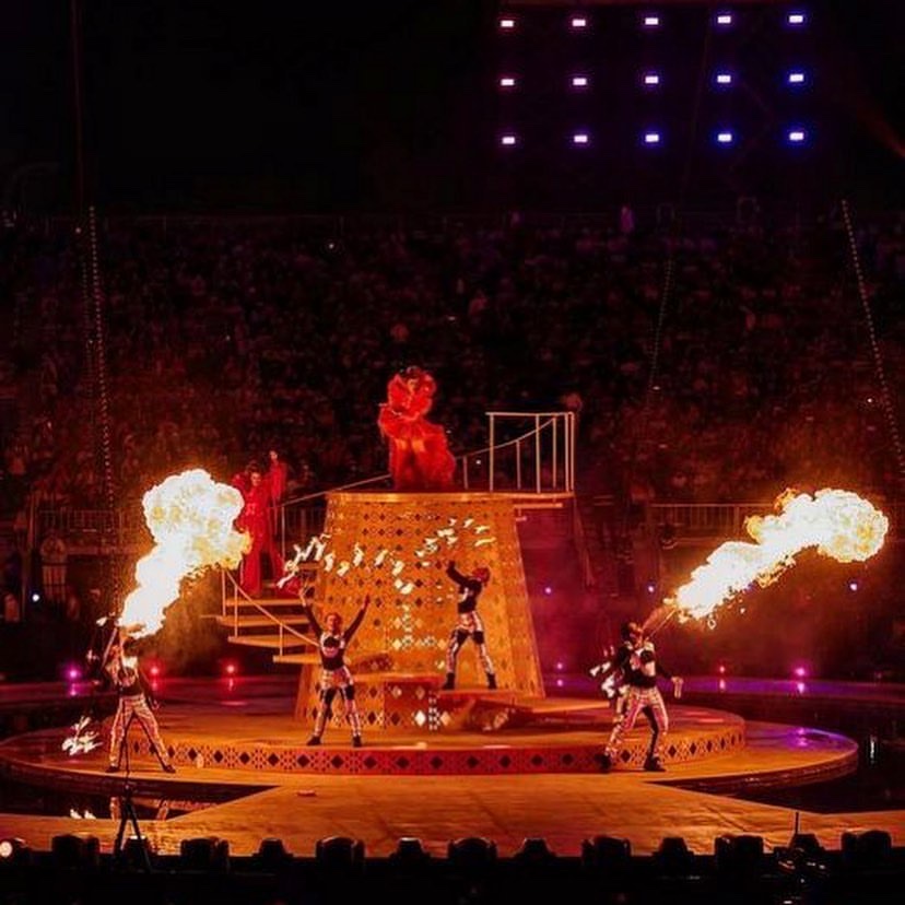 Performers on and around a tall golden set piece in a stadium, flame jets firing on either side.