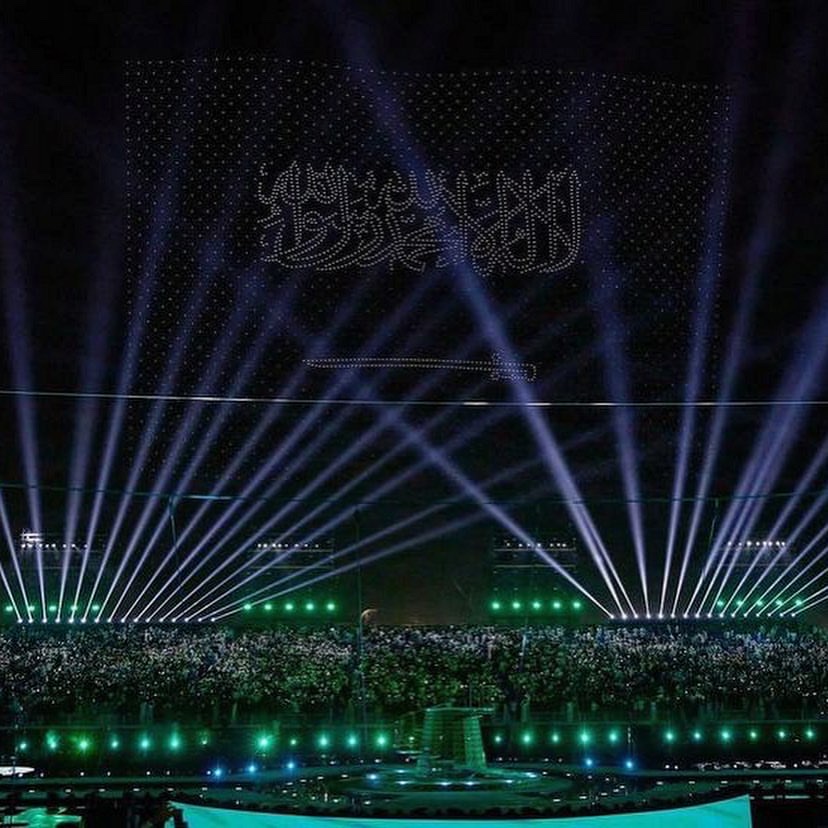 A drone formation spelling Arabic script above a stadium, searchlight beams fanning up from the floor.