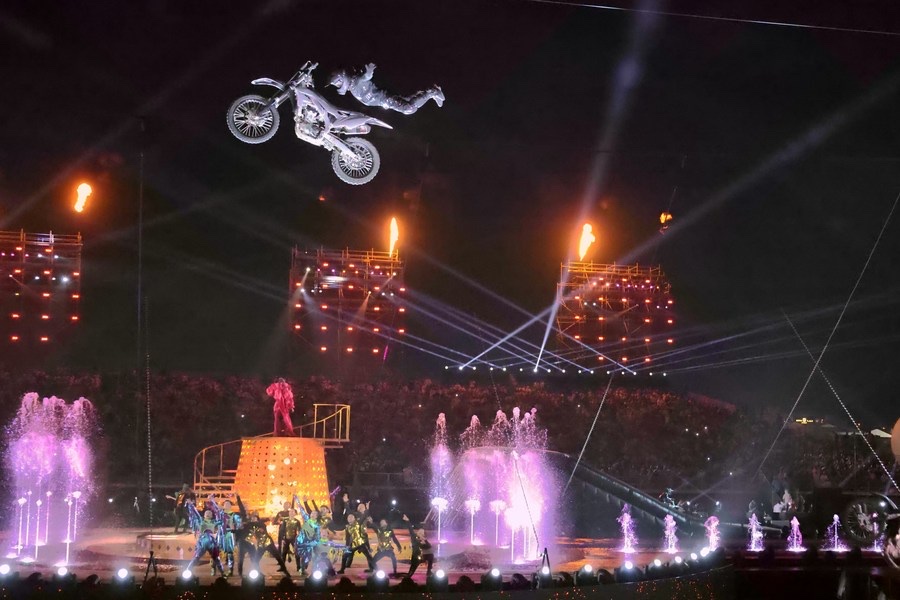 A motorcycle rider in mid-air above a stadium arena, water jets and performers on the ground below.