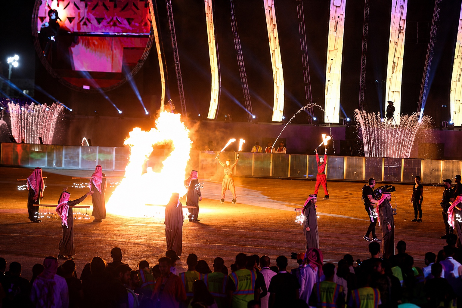 Performers with flame torches around a burning shape on the arena floor, spectators watching from the barrier.