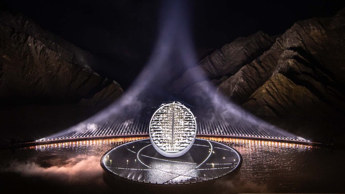 A large illuminated ovoid sculpture on a circular stage in a mountain amphitheatre, lit from above through mist.