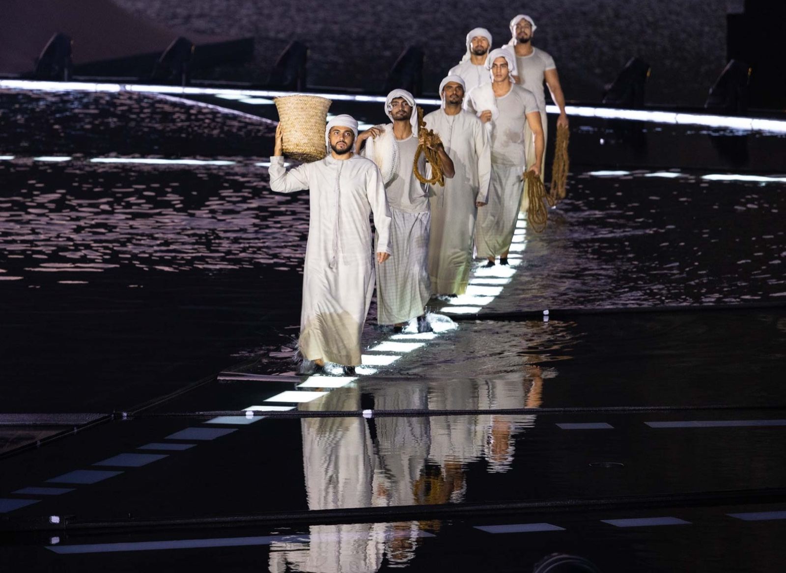 Performers in white robes walking in single file along a lit walkway, the leading performer carrying a woven basket.