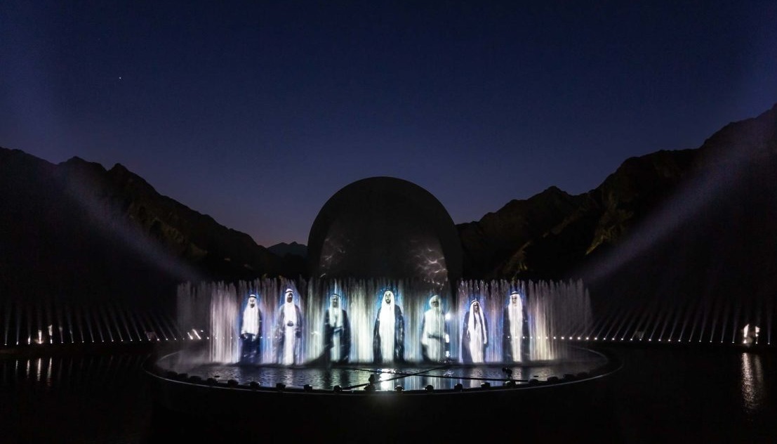 A row of performers standing between tall water jets in front of the domed set piece.
