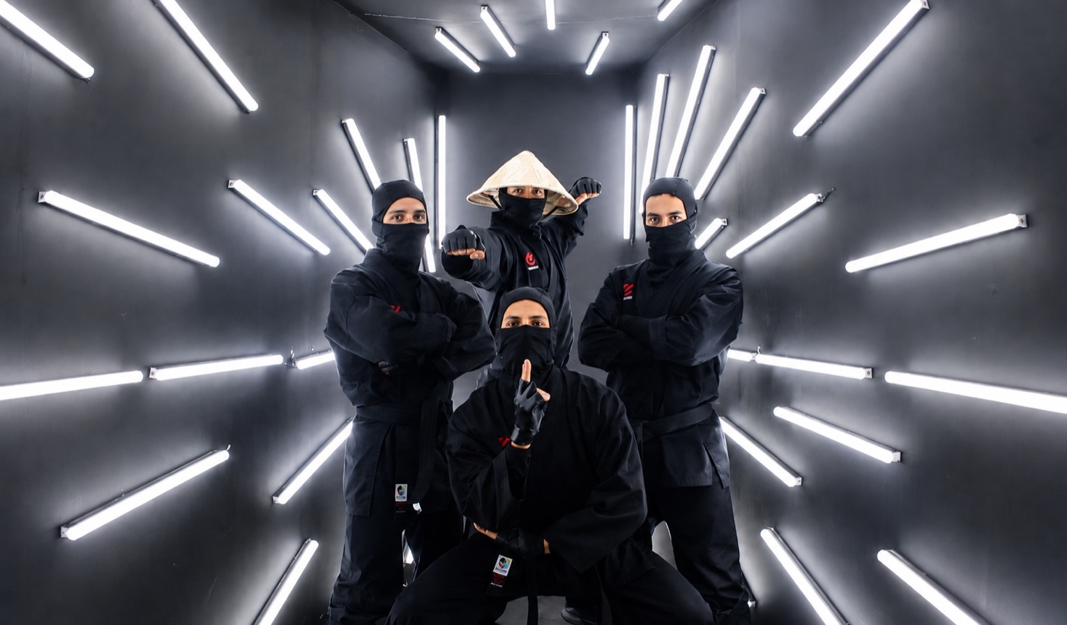 Four performers in black ninja costume posed in a corridor lined with angled white light tubes.