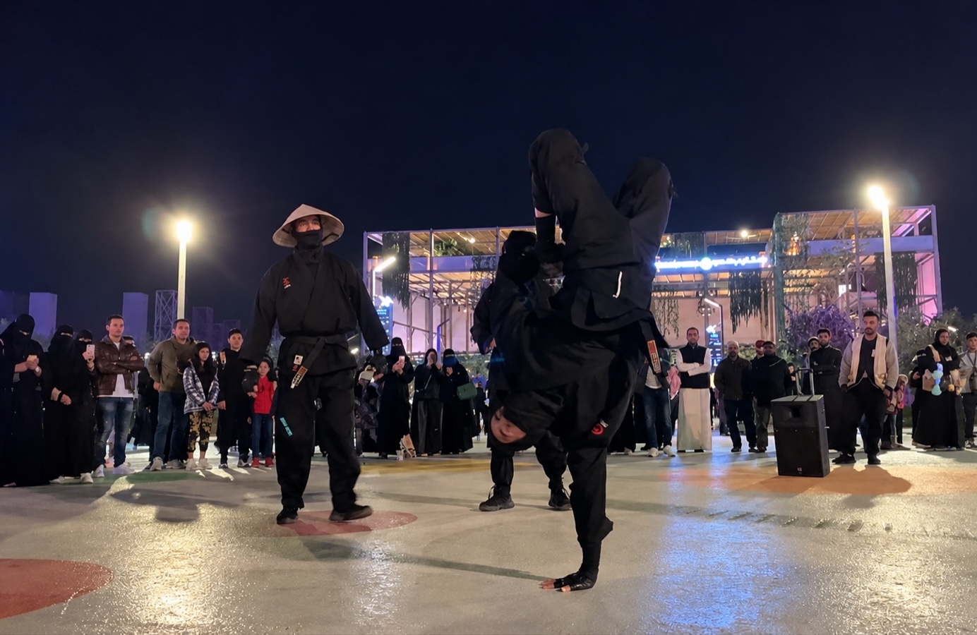 Performers in ninja costume in an outdoor plaza at night, one balanced upside down, onlookers gathered around.