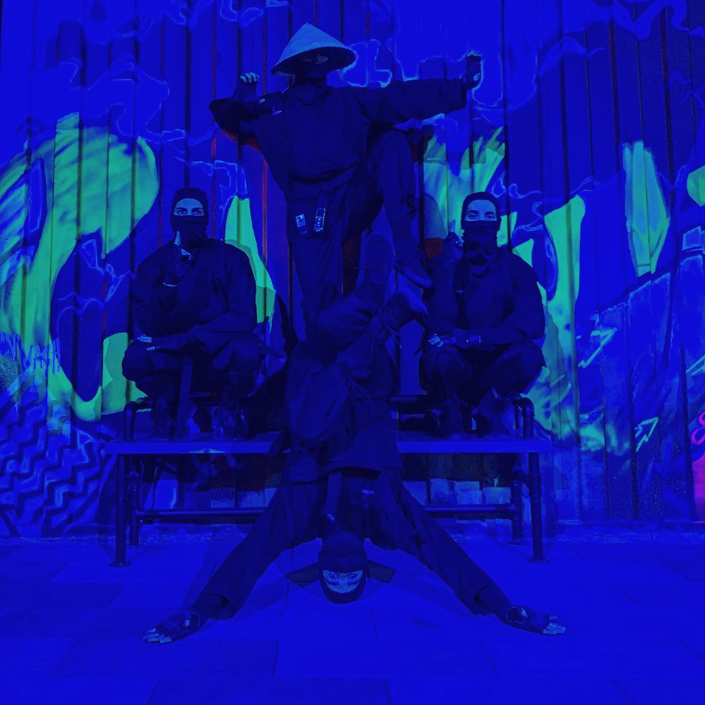 Performers posed on and around a bench against a graffiti wall, the whole scene lit deep blue.