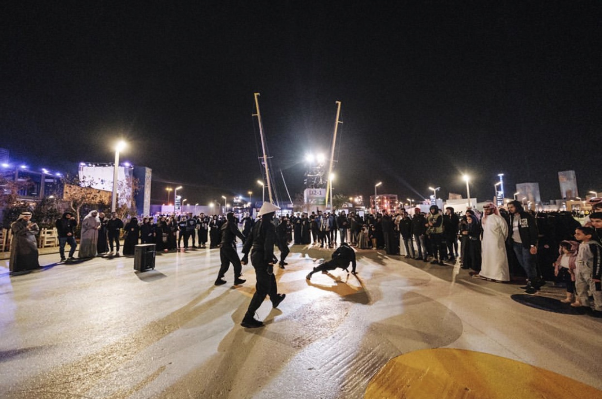 Wide night view of an outdoor performance, one performer low to the ground as a crowd watches from both sides.