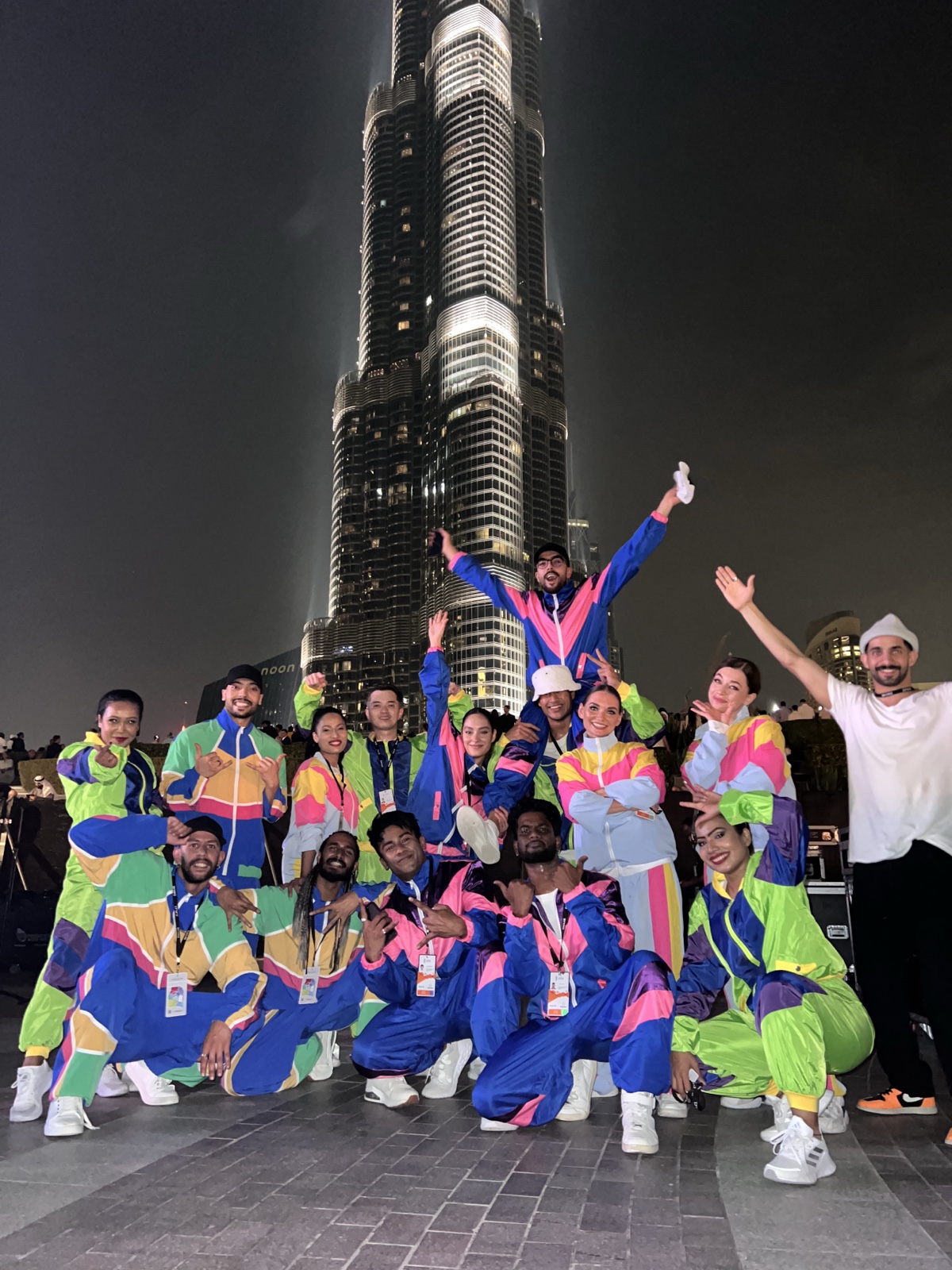 The cast in bright colour-blocked tracksuits grouped together in front of the Burj Khalifa at night.