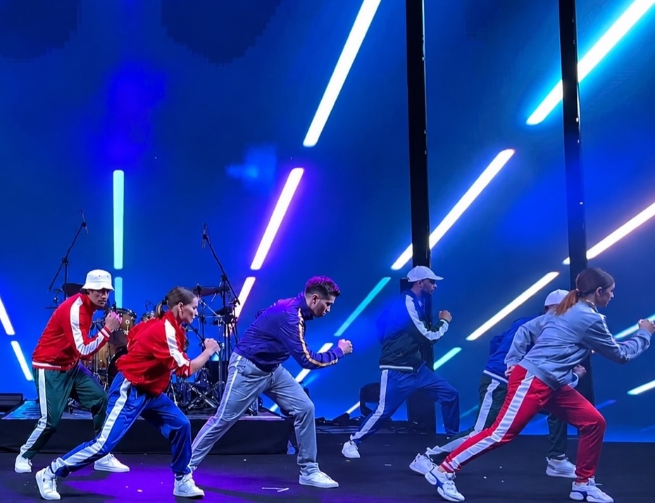 Dancers in red, blue and grey tracksuits mid-routine on stage against a backdrop of blue neon bars.
