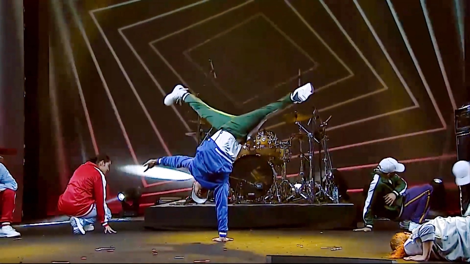 A dancer holding a one-handed freeze in front of a drum kit, two others crouched to either side.
