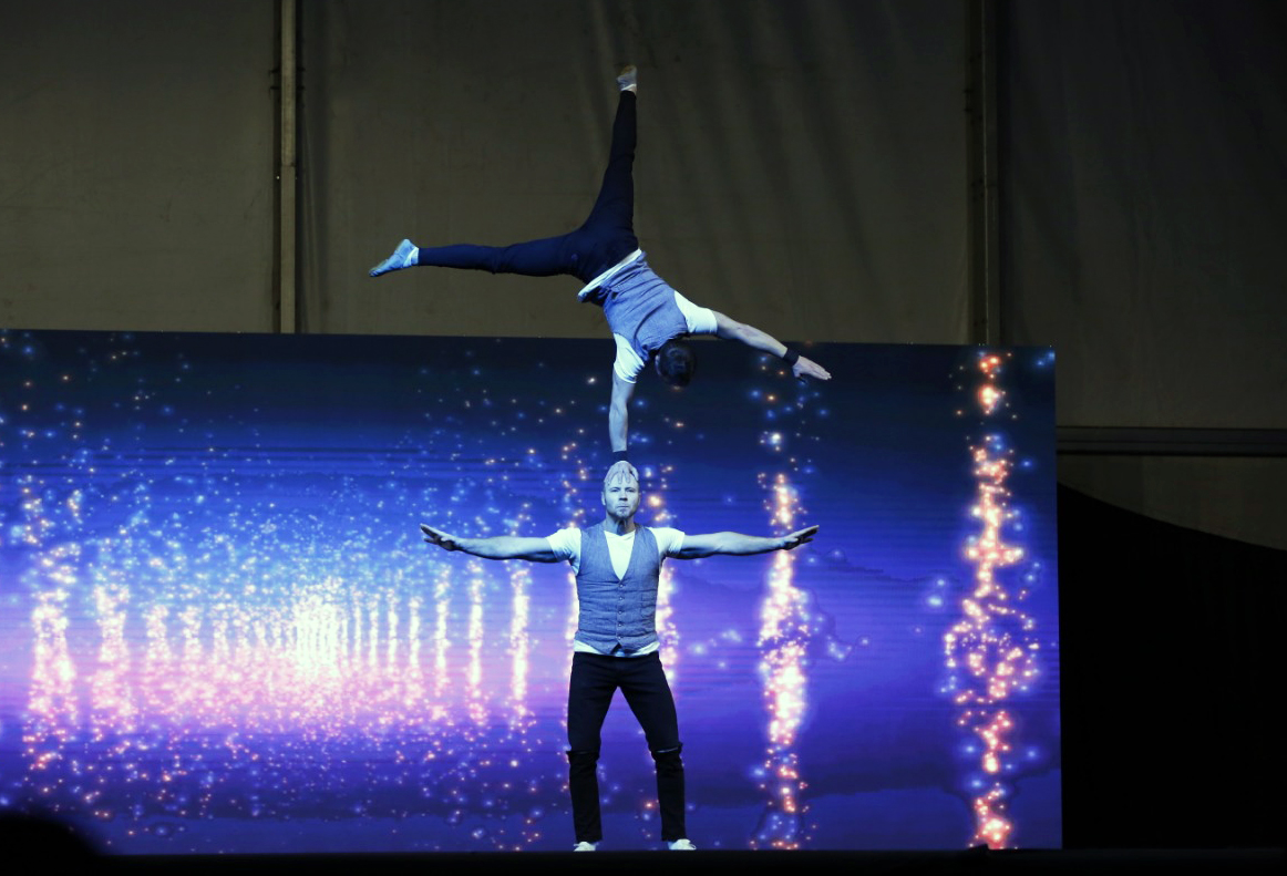 An acrobat in a one-arm handstand balanced on a partner's head, in front of a screen of blue sparks.