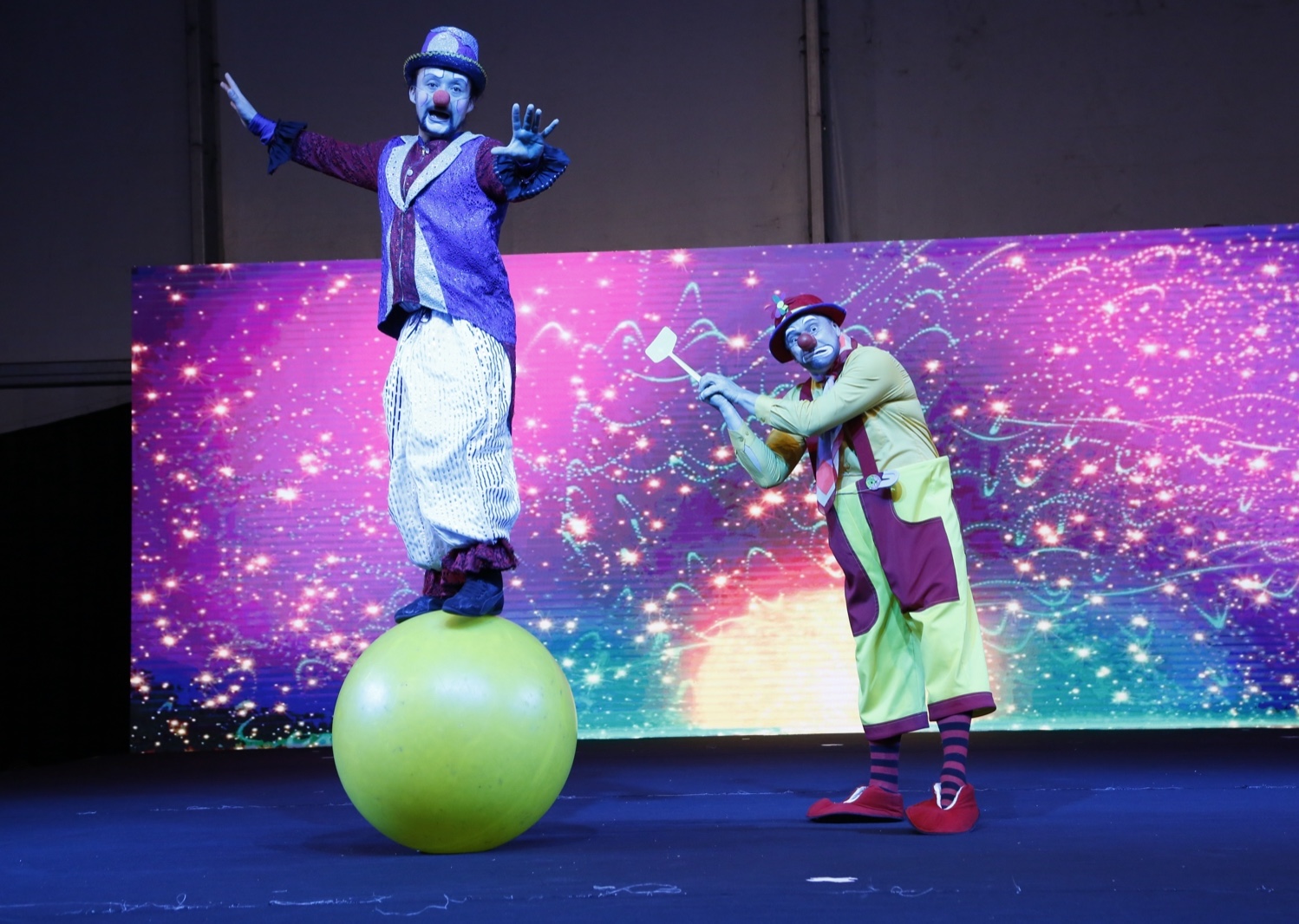Two clowns performing, one balanced standing on a large green ball, against a bright magenta screen.