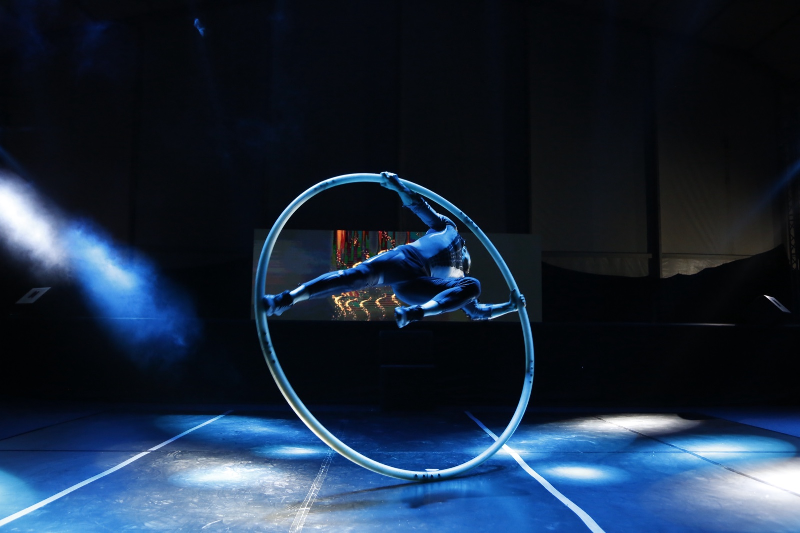 A performer inside a Cyr wheel, tilted mid-spin under crossed blue beams.