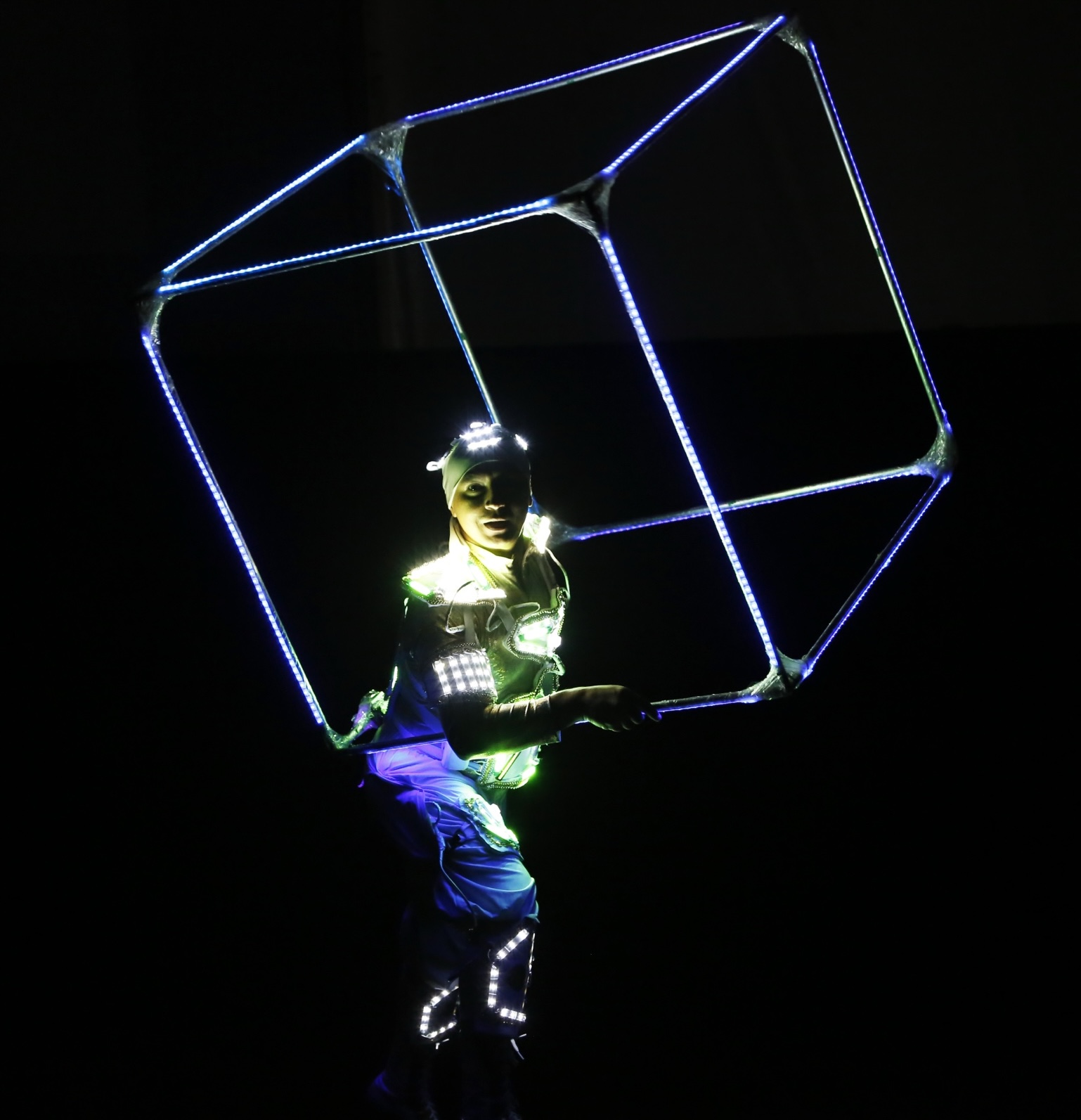 A performer in an LED costume holding a large illuminated cube frame around the body.