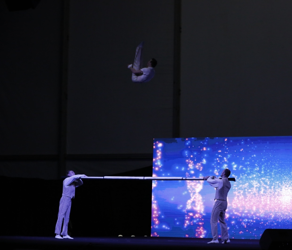 Two performers brace a long flexible bar while a third somersaults high above it.