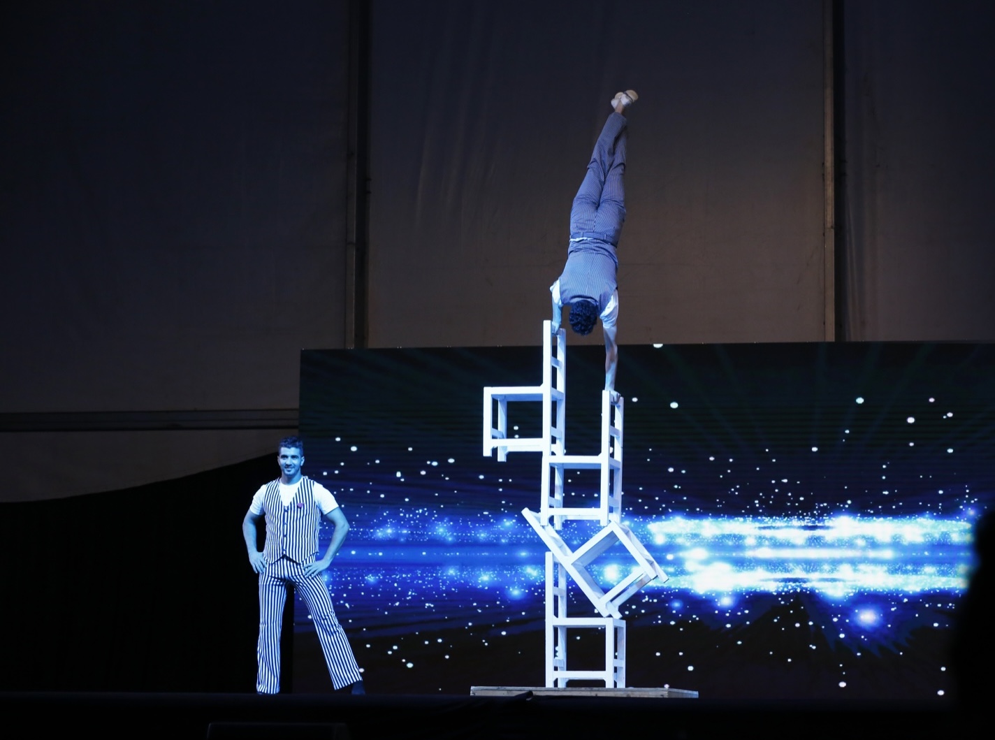 An acrobat holding a handstand on a tower of stacked chairs as another performer watches from the stage.