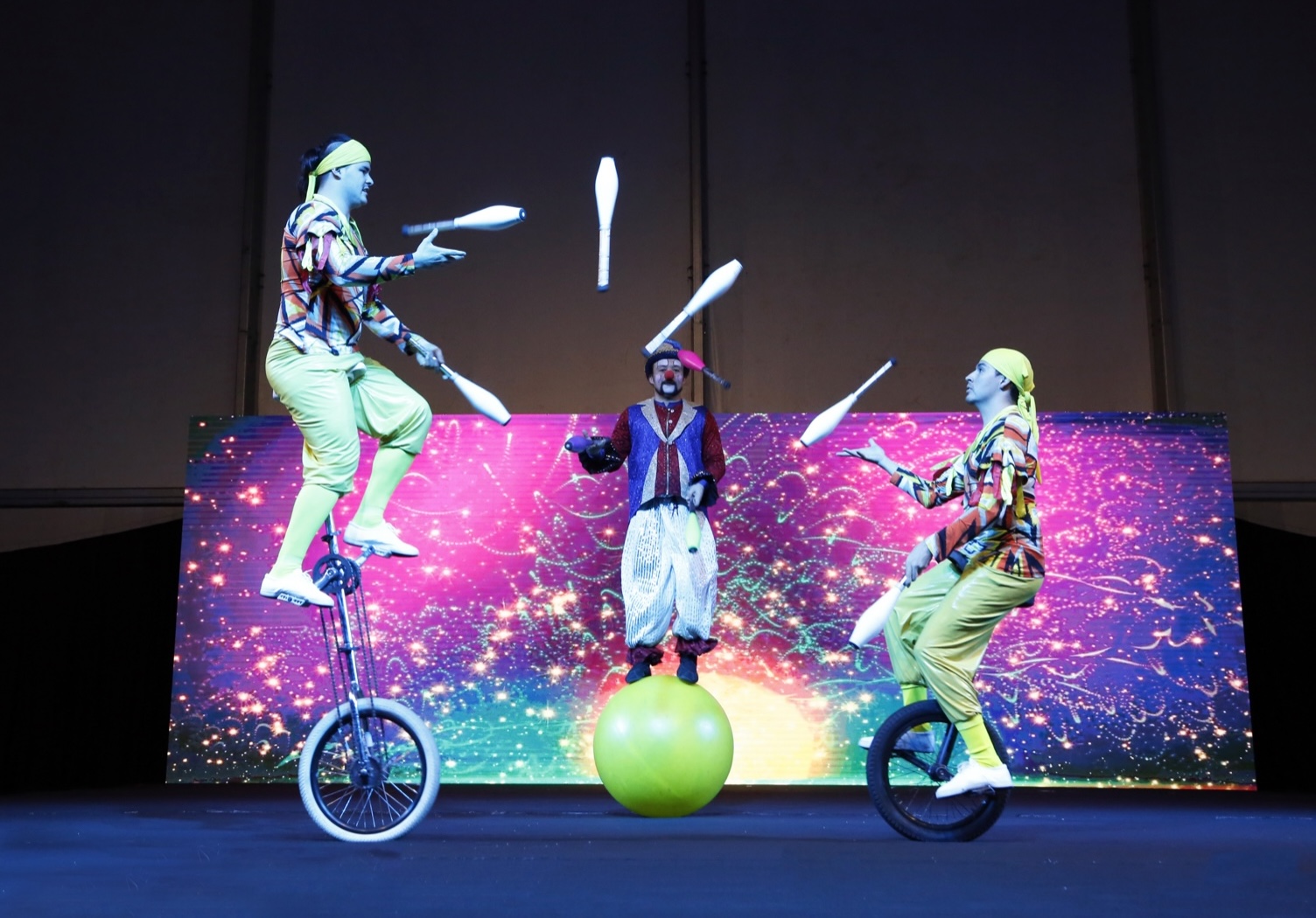 Three clowns juggling together, two on unicycles and one standing on a large green ball.