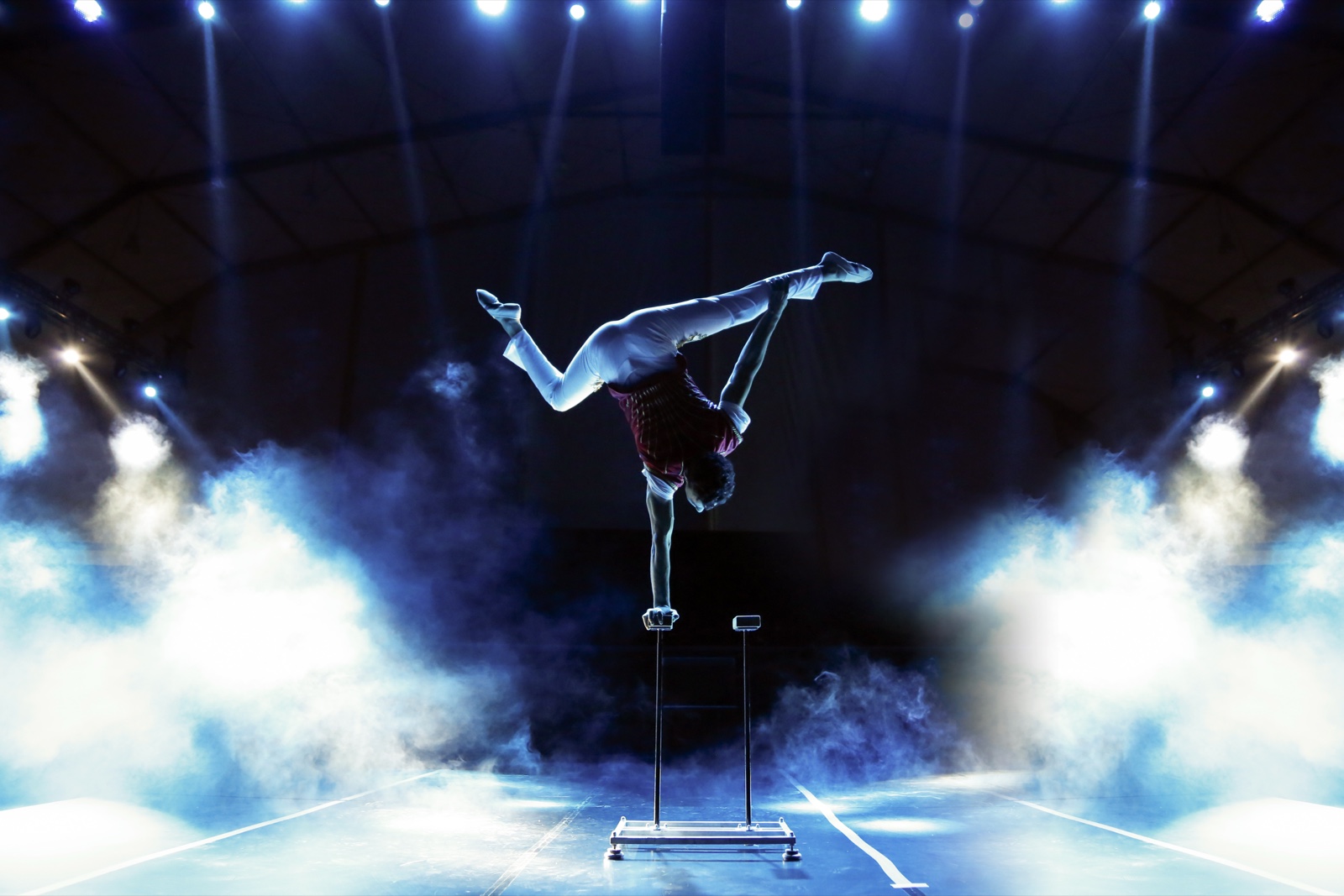 A performer holding a one-arm handstand on canes, lit through haze by crossed beams.