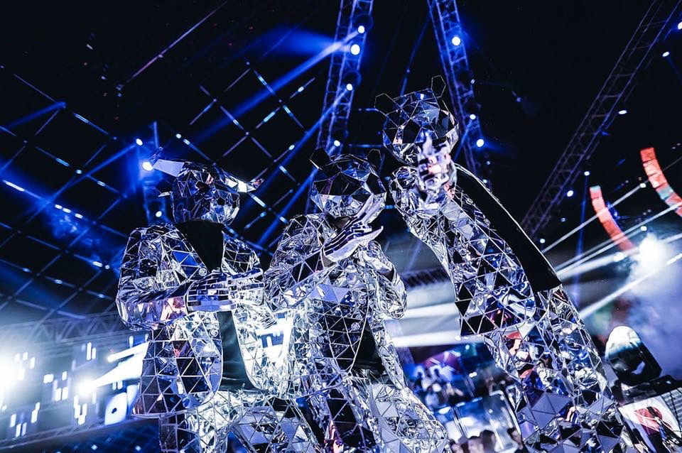 Three performers in mirrored mosaic costumes catching the blue stage lighting above a crowd.