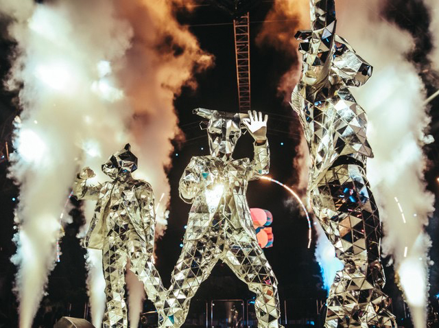 Mirrored-costume performers on stage flanked by tall white pyrotechnic fountains.
