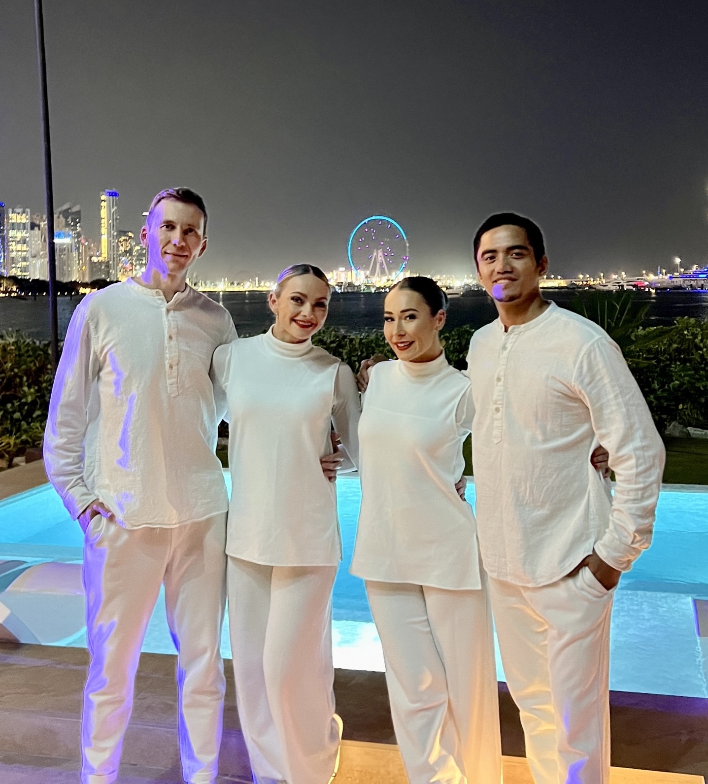 Four performers dressed in white beside a rooftop pool at night, the Dubai skyline and Ain Dubai behind them.