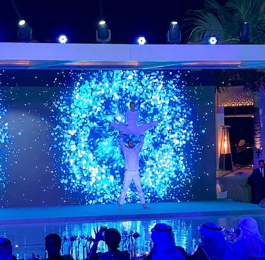 An acrobat lifted overhead by a partner in front of a screen showing a burst of blue particles, guests watching from tables.