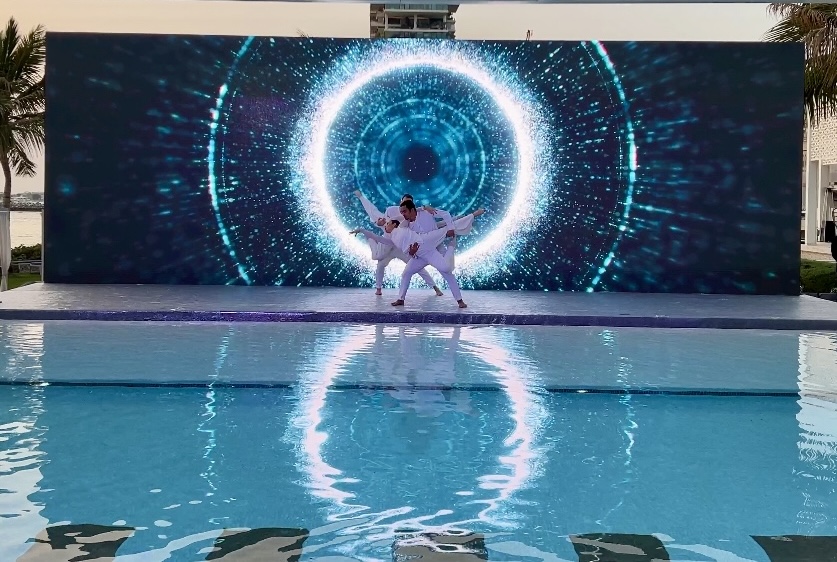 Two performers posed in front of a screen showing a bright blue vortex, mirrored in the pool below.