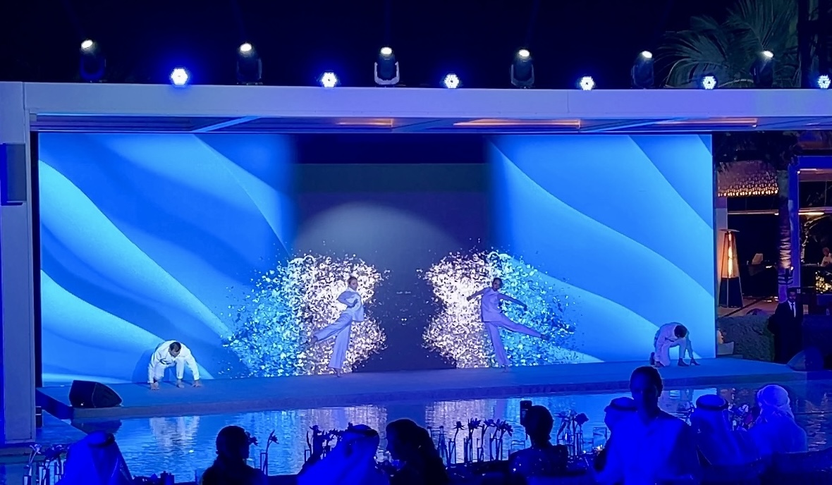 Wide view of the poolside stage: dancers in white against a deep blue screen, guests seated around the water.